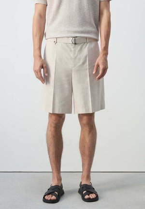 Hombre que lleva pantalones cortos plisados beige con cinturón y sandalias negras, de pie frente a un fondo claro y liso.