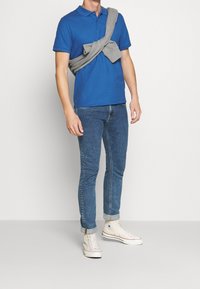 Polo bleu, pull gris posé sur les épaules, jeans denim slim et baskets blanches montantes. Design uni, sans motifs visibles.