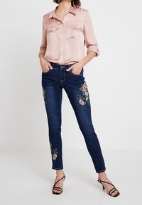 Ljusrosa satinsskjorta med två bröstfickor, kombinerad med mörkblå jeans med blommönster på framsidan. Svarta remsandaler.