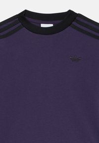 Camiseta de algodón morada con cuello redondo acanalado negro, que presenta tres rayas negras en los hombros y un detalle de logo negro en el pecho.
