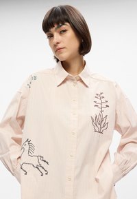 Ανοιχτό ροζ καρό πουκάμισο με μακριά μανίκια, κεντημένες σχέσεις μονόκερου και λουλουδιών, κούμπωμα με κουμπιά και μαλακή βαμβακερή υφή.