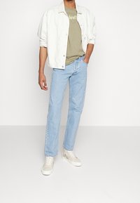 Muž v světlých modrých džínách, béžových grafických vysokých teniskách, olivově zeleném tričku Levi's a bílé košili s vyhrnutými rukávy.