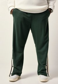 Dark green sweatpants with side snap details, featuring white accents and pockets. Soft texture, relaxed fit, and tapered legs.