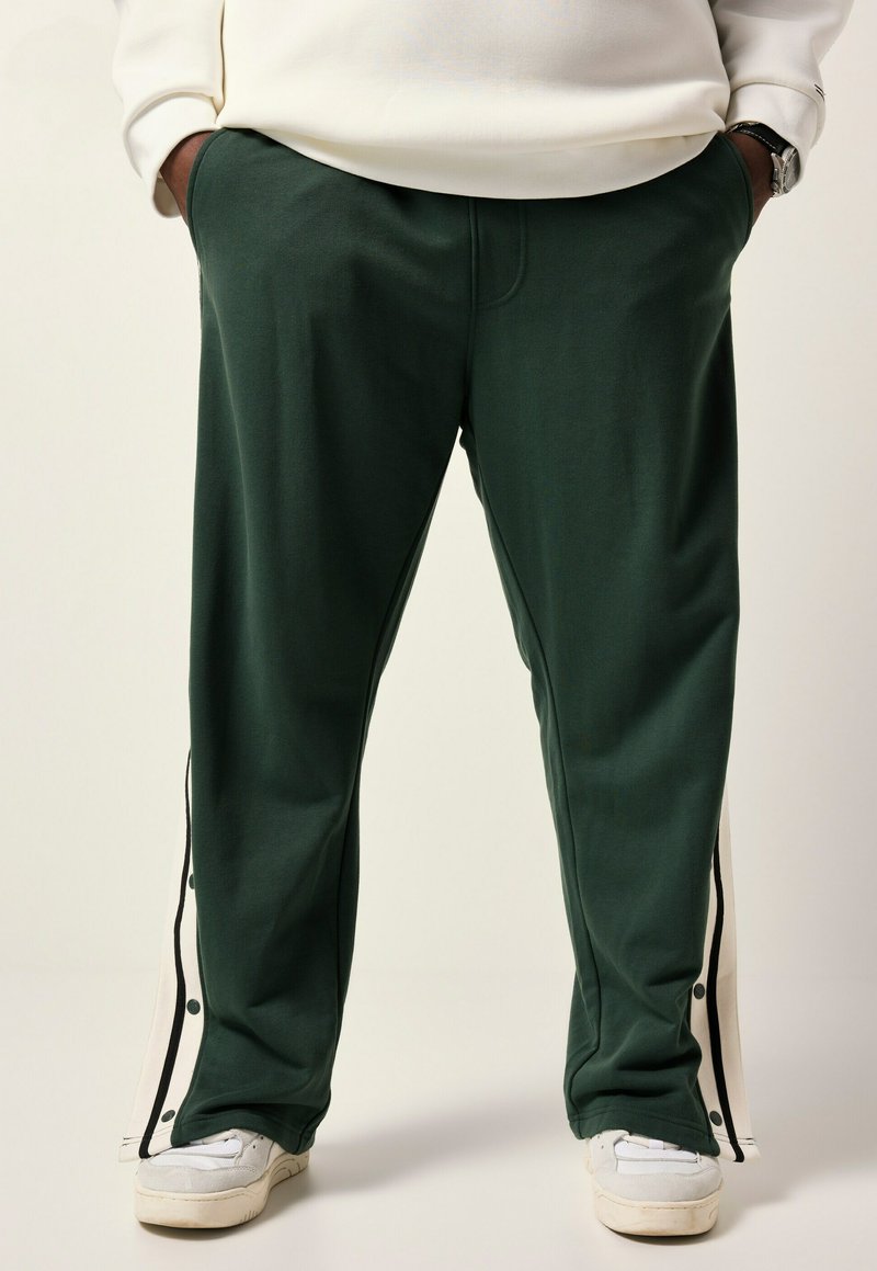 Dark green sweatpants with side snap details, featuring white accents and pockets. Soft texture, relaxed fit, and tapered legs.