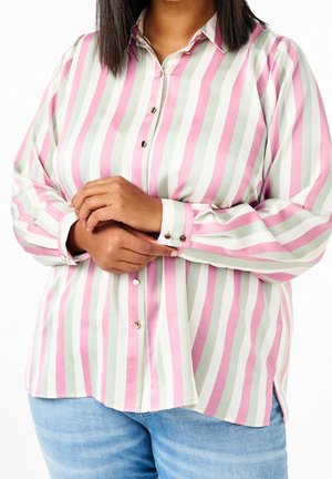 Kvinde iført en pink, hvid og grå vertikalt stribet skjorte med manchetknapper, der justerer den ene manchet, kombineret med lyseblå jeans.