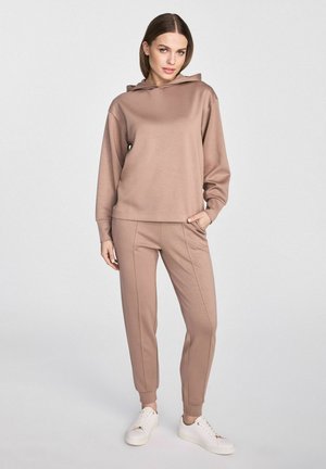 Jasnobrązowy dres z hoodie i joggerami wykonany z miękkiego materiału. Hoodie ma kieszeń z przodu, luźny krój i ściągacze przy mankietach. Białe sneakersy dopełniają stylizację.