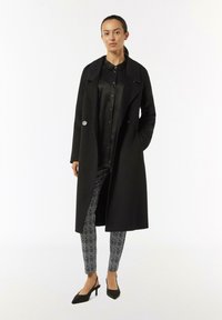 Schwarzer, langer, taillierter Mantel mit doppelt geknöpften Knöpfen, getragen über einer schwarzen Bluse und gemusterten Hosen. Spitz zulaufende schwarze Absätze runden das Outfit ab.