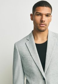 Junger Mann mit kurzem Haar und gestutztem Bart, der ein hellgraues Blazerjackett über einem schwarzen gerippten Shirt trägt und direkt in die Kamera schaut, vor einem schlichten Hintergrund.