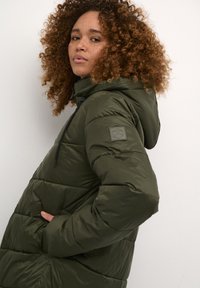Olive green puffer jacket with a hood, featuring a quilted design, side pockets, and a logo patch on the left sleeve. Smooth texture.