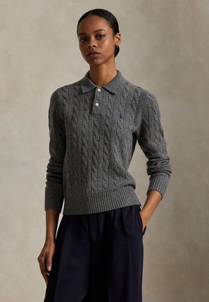 Femme aux cheveux foncés coiffés en chignon bas, portant un pull polo gris en maille torsadée à manches longues et un pantalon bleu marine foncé, debout avec une main dans la poche.