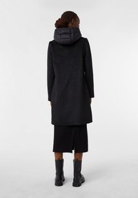 Cappotto nero con cappuccio trapuntato, maniche lunghe e taglio dritto, abbinato a una gonna nera e stivali neri alla caviglia.