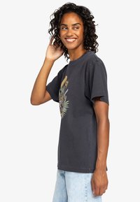 T-shirt nera in cotone con un design grafico colorato che presenta elementi tropicali. Maniche corte e vestibilità rilassata, abbinata a jeans blu chiaro.