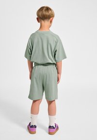 Ljust grönt avslappnat t-shirt och shorts i mjukt tyg, med elastisk midja. Barnet har på sig vita strumpor och lila skor.