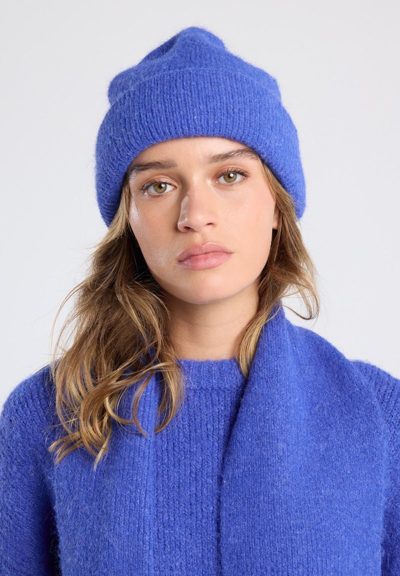 Blauwe gebreide beanie met geribbelde textuur, gemaakt van zacht materiaal. Gecombineerd met een bijpassende trui, die een samenhangende kleur en ontwerp laat zien.