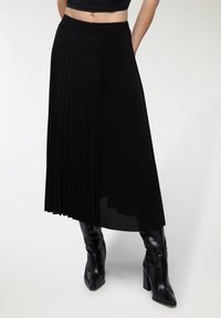 Gonna midi plissettata nera con vita liscia e orlo trasparente a contrasto, abbinata a stivali neri con tacco aderenti.