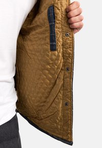 Quilted brown jacket interior with a textured finish; features a denim pocket and snap buttons along the edge.