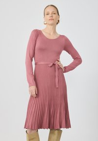 Ribbet, langermet kjole i myk rosa med tettsittende liv, belte i midjen og plisserte skjørt. Glatt tekstur med en flytende silhuett.