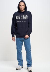 Tamsiai mėlynas sukapotas džemperis su baltu užrašu "BIG STAR" ir kišene, derinamas su šviesiai mėlynomis džinsomis ir pilkais bei baltais sportbačiais.