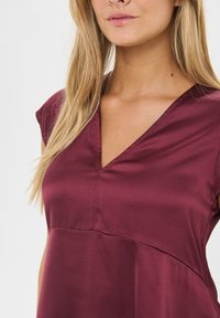 Burgundy klänning med V-ringning och korta ärmar, tillverkad av ett slätt tyg, med en söm detajl över bröstet och en flödande silhuett.