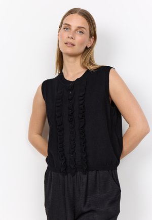 Woman with light hair wears a sleeveless black top with vertical ruffles and buttons, paired with loose black pants against a white background.