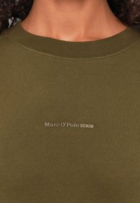 Alyvuogių spalvos medvilninė megzta džemperis su apykakle, turintis įrengtą apykaklę ir atspausdintą "Marc O'Polo DENIM" logotipą ant krūtinės. Minkšta tekstūra ir paprastas dizainas.