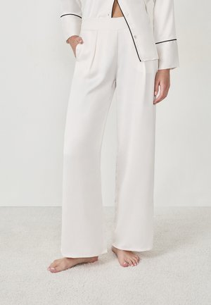 Femme portant un pantalon en satin blanc, ample et taille haute, avec des plis, se tenant pieds nus sur un tapis clair devant un fond blanc uni.