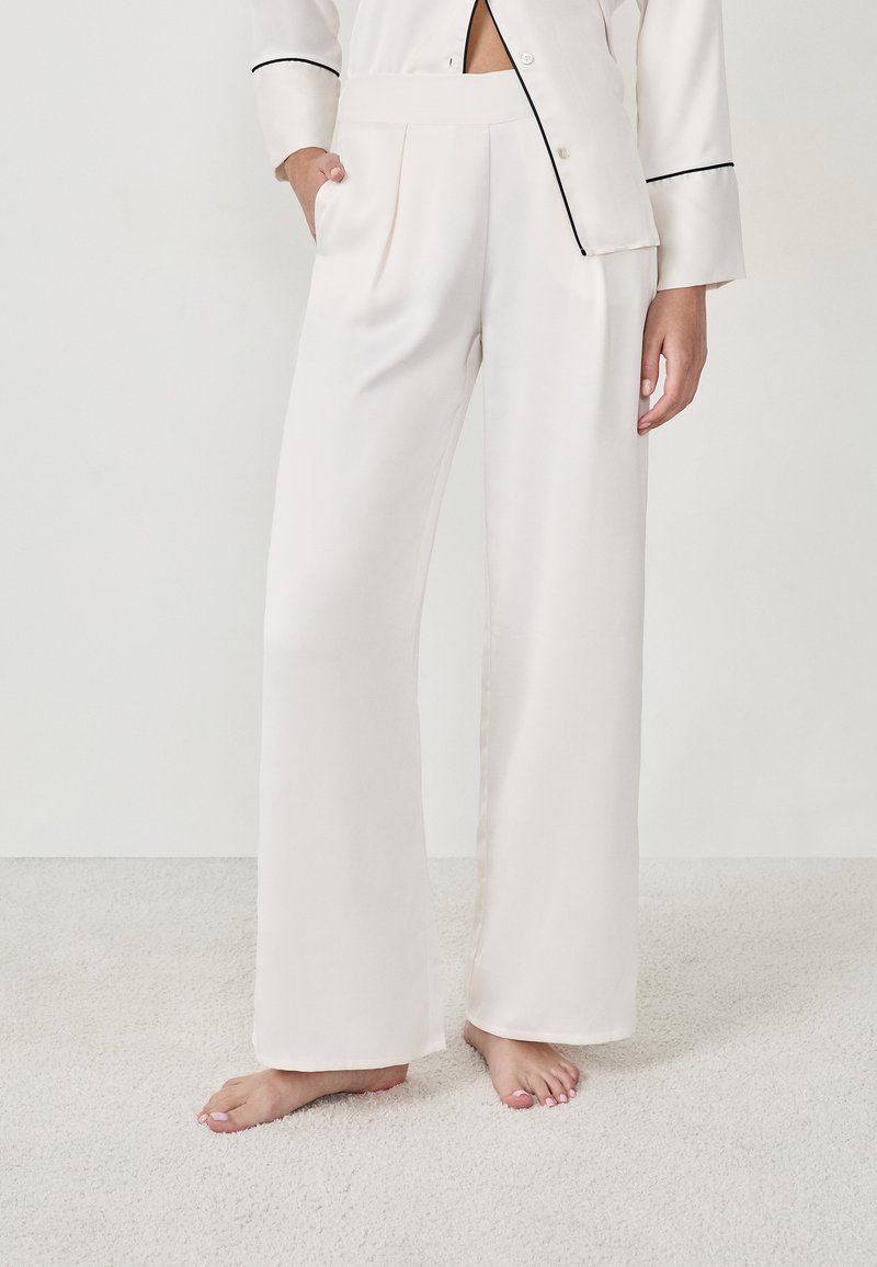 Femme portant un pantalon en satin blanc, ample et taille haute, avec des plis, se tenant pieds nus sur un tapis clair devant un fond blanc uni.