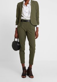 Mujer que lleva un traje verde oliva con cinturón negro, camisa blanca de botones, sandalias de tacón alto negras con tiras, sosteniendo un bolso redondo negro.