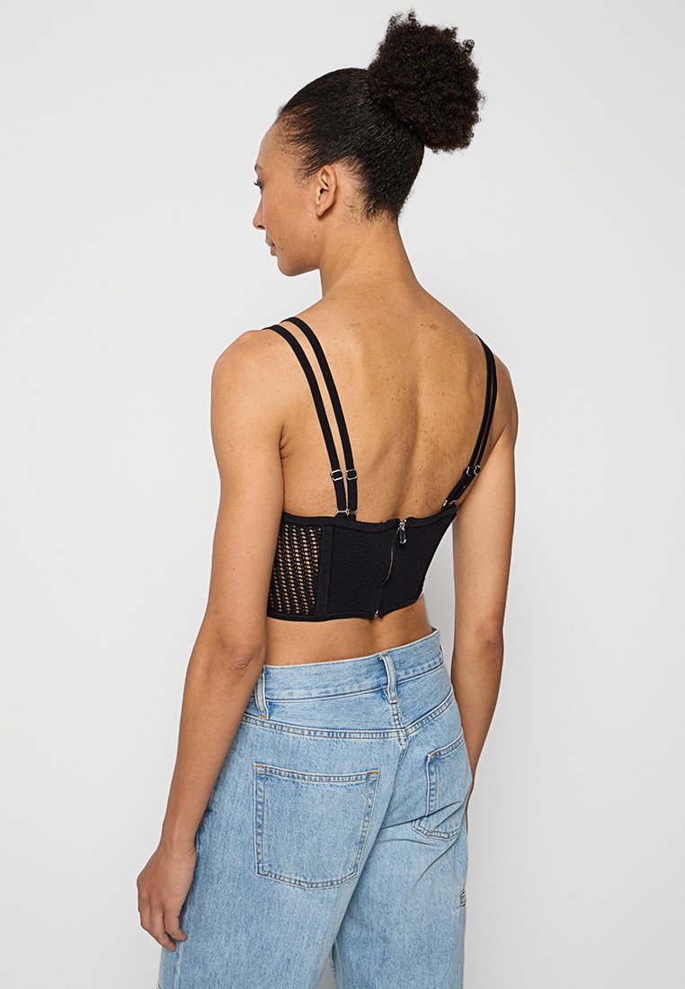 Schwarzes Mesh-Korsett-Top mit verstellbaren Trägern, das einen Reißverschluss auf der Rückseite und ein strukturiertes Strickdesign aufweist. Kombiniert mit hellblauen Jeans.