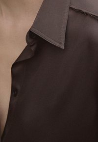Brun button-up skjorte med en struktureret krave, glat tekstur og en synlig sømdetalje langs kanten af kraven og knappene.
