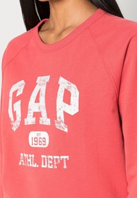 Felpa color corallo con scollo rotondo e maniche raglan. Presenta testo bianco consumato con scritto "GAP" e "ATHL DEPT," insieme a una data di fondazione.