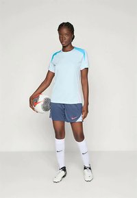 Sieviete gaiši zila sporta krekla, tumši zilu šortos, baltās zeķēs un sporta apavos, turot futbola bumbu, stāvot pret vienkāršu fona.