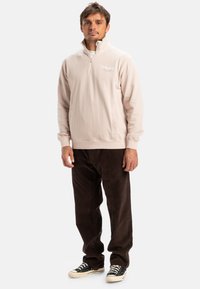 Light pink zip-up sweatshirt with a high collar, side pockets, and a subtle logo. Paired with dark brown corduroy pants and black sneakers.