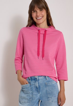 Femme souriante portant un haut à manches longues rose à motifs avec des cordons et un jean bleu clair avec de grandes poches avant, main dans la poche.