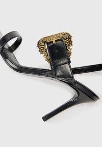 Black high-heeled sandals with thin straps, featuring a gold ornate buckle. The design includes a smooth leather texture and pointed toe.