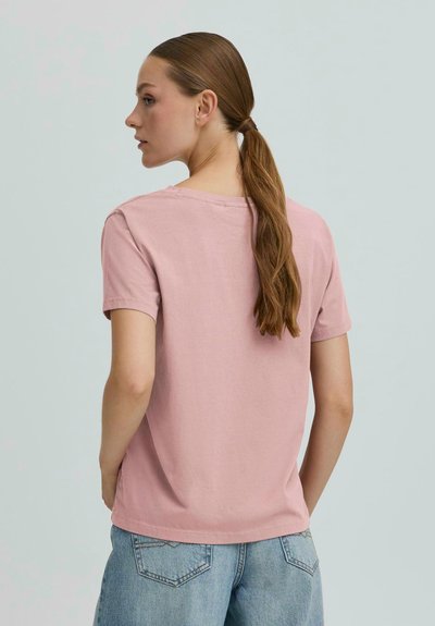 Žena s dlhým hnedým copom, oblečená v ružovom tričku s krátkym rukávom a svetlomodrých džínsoch, stojí bokom na jednofarebnom pozadí.