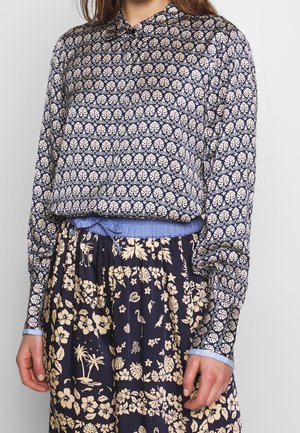 Femme portant un chemisier bleu marine à manches longues avec des motifs de feuilles crème et une jupe bleu marine avec des imprimés floraux tropicaux et oiseaux crème.