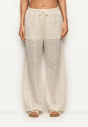 Kobieta ubrana w kremowe, szydełkowane spodnie z wysokim stanem i szerokimi nogawkami, z regulowanym sznurkiem w talii oraz pasujące sandały z odkrytymi palcami, stojąca na jednolitym białym tle.