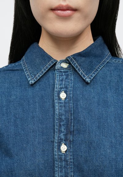 Τζιν πουκάμισο με κλασικό γιακά, πέντε λευκά πλαστικά κουμπιά και μεσαίο μπλε χρώμα με διακριτική υφή και λεπτομέρειες ραφής.