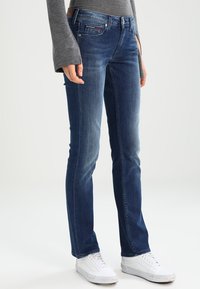 Vaqueros de mezclilla azul con un diseño de pierna recta, estilo de cinco bolsillos y un ligero desvanecimiento. Combinados con zapatillas blancas y una camiseta de manga larga gris.