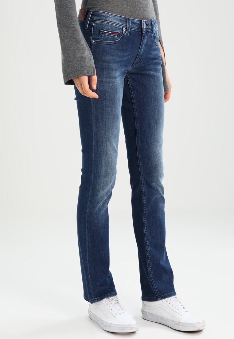 Vaqueros de mezclilla azul con un diseño de pierna recta, estilo de cinco bolsillos y un ligero desvanecimiento. Combinados con zapatillas blancas y una camiseta de manga larga gris.