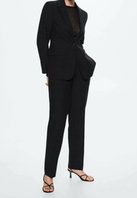 Sort skræddersyet jakkesæt med en enkelt knap-blazer, lige ben-bukser og en gennemsigtig sort top. Beklædningen har et minimalistisk design og glatte linjer.