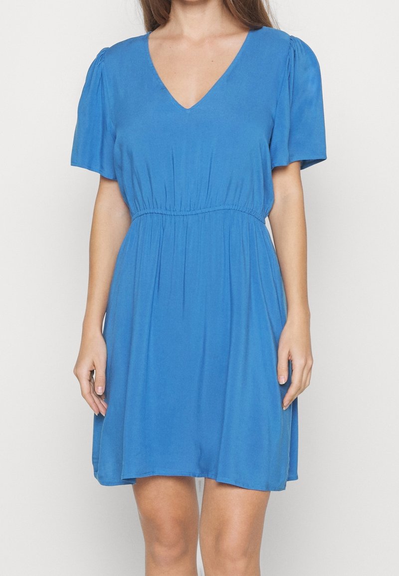 Femme portant une robe bleue à manches courtes avec un col en V et une taille froncée, debout devant un fond clair uni.