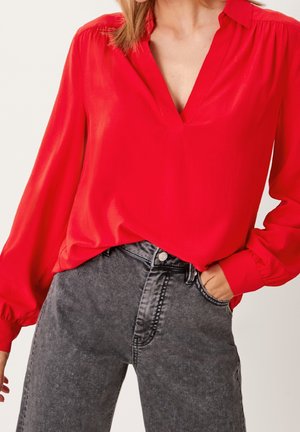Kvinde iført en skarp rød bluse med lange ærmer og V-hals samt højttaljede, slidte sorte jeans, med den ene hånd i lomma.