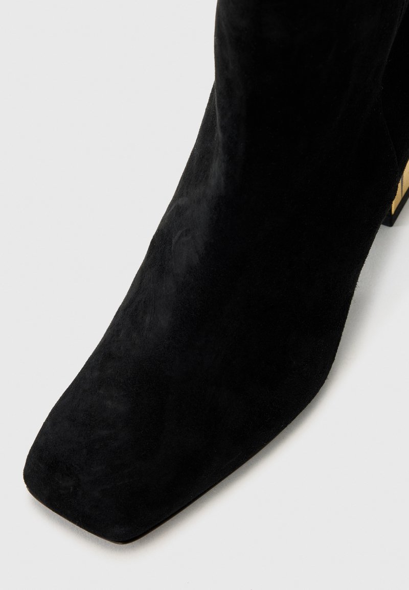 Botte de cheville en suede noire avec un bout carré et un talon épais ; texture lisse avec un design minimaliste et sans embellissements visibles.