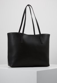 Bolso tote de cuero negro texturizado con dos correas largas para el hombro, exhibido sobre un pedestal blanco en un fondo claro y liso.