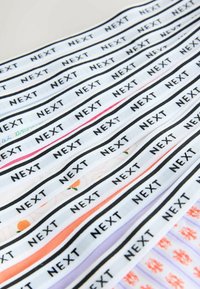 Bandes de tissu blanc avec le mot "NEXT" imprimé en noir de manière répétée, disposées en diagonale sur des textiles à motifs en arrière-plan.