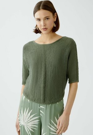 Green knitted sweater with a wide neckline and short sleeves, featuring a textured panel down the center. Worn over patterned light green pants.