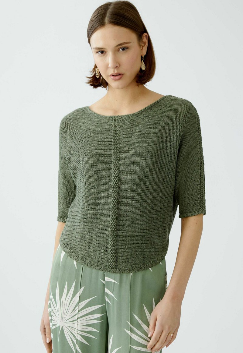 Grüner Strickpullover mit weitem Halsausschnitt und kurzen Ärmeln, der ein strukturiertes Panel in der Mitte aufweist. Über gemusterten hellgrünen Hosen getragen.