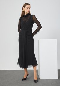 Μαύρο φόρεμα με διάφανες μακρύες μανίκες και συγκεντρωμένη μέση. Διαθέτει ρέουσα σιλουέτα και μαλακό, ελαφρύ ύφασμα. Συνδυάζεται με μαύρες γόβες.